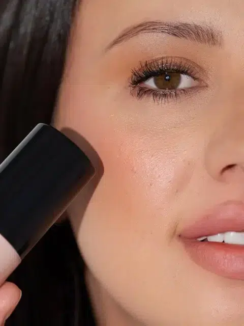 Close-up of a person holding a makeup product next to their face, showcasing a smooth complexion with a hint of highlighter.