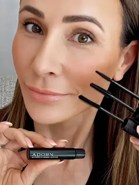 A person holding a small Adorn makeup product near their face while also holding two mascara wands in the other hand, with a neutral expression.