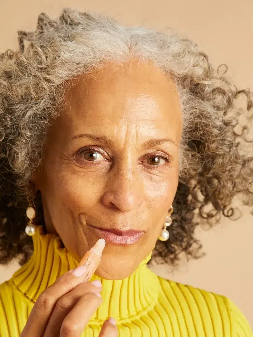 A woman with curly gray hair and a yellow turtleneck applies lip balm to her lips. She has pearl earrings and is smiling slightly.