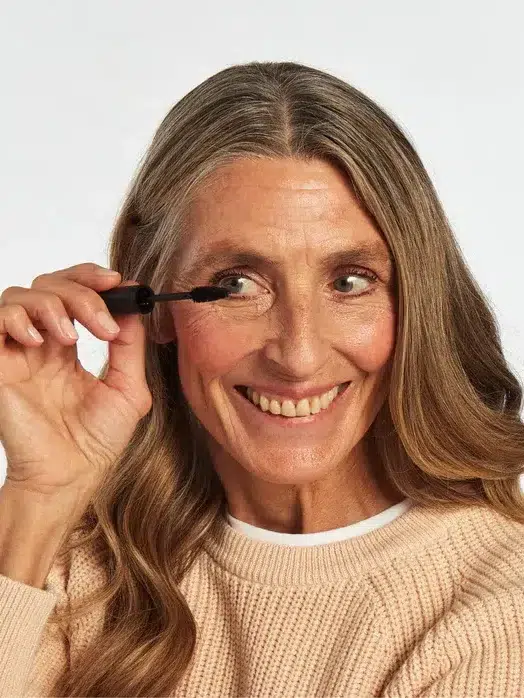 A woman with long hair smiles while applying mascara to her eyelashes. She is wearing a beige sweater and a white shirt, and the background is plain white.