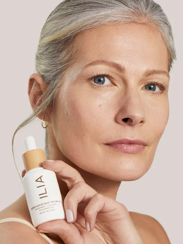 A woman with light skin and gray hair holding a bottle of ILIA Super Serum Skin Tint SPF 40 near her face.