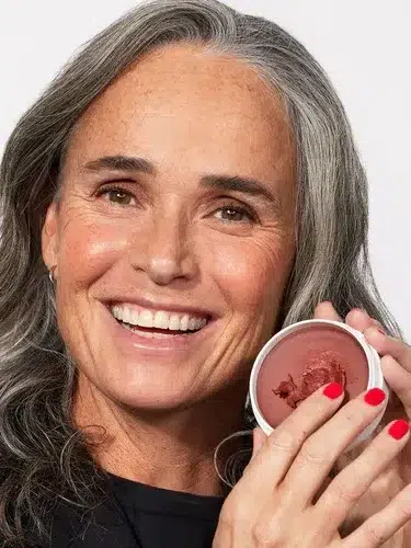 A smiling person with gray hair holds an open container with a reddish-brown product. Their nails are painted red.