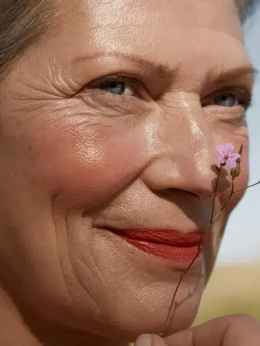 An older woman with red lipstick holds a small pink flower close to her face, smiling softly.