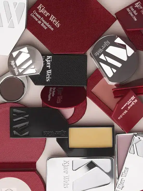 A variety of Kjaer Weis cosmetic products are arranged on a surface, including compacts, containers, and palettes with red and silver packaging.