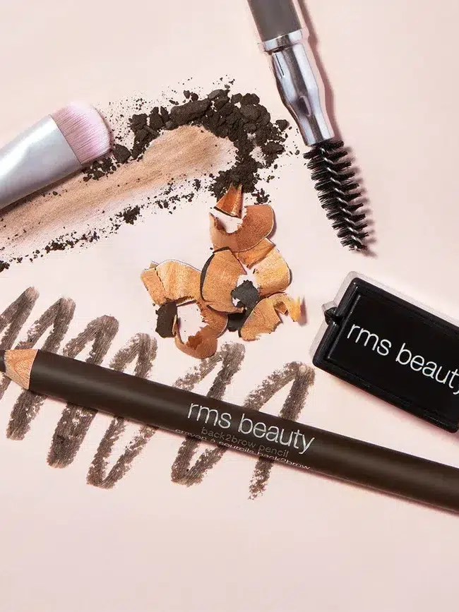 Close-up of RMS Beauty eyebrow pencil, sharpener, and brush, with shavings and swatches on a light background.