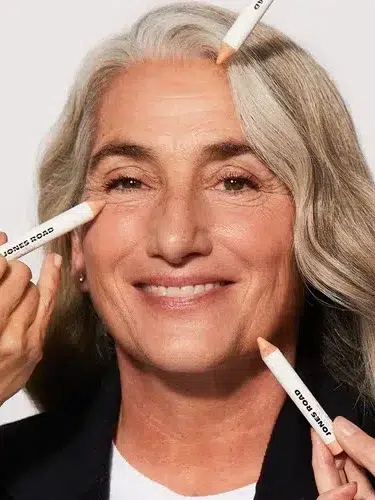 A smiling person with gray hair is having concealer applied under the eyes and on the cheekbones by multiple hands holding concealer sticks.