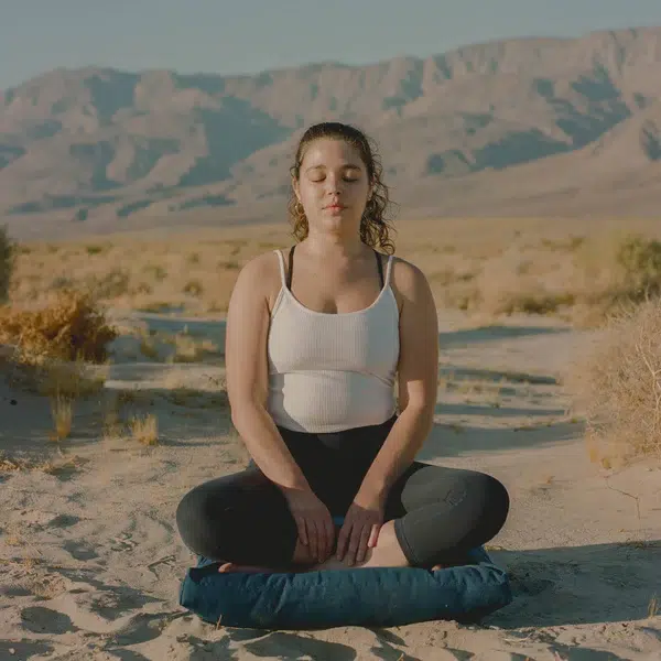 Person meditating in a desert landscape, sitting cross-legged on a cushion, with eyes closed and hands resting on their knees. Mountains and shrubs are visible in the background.