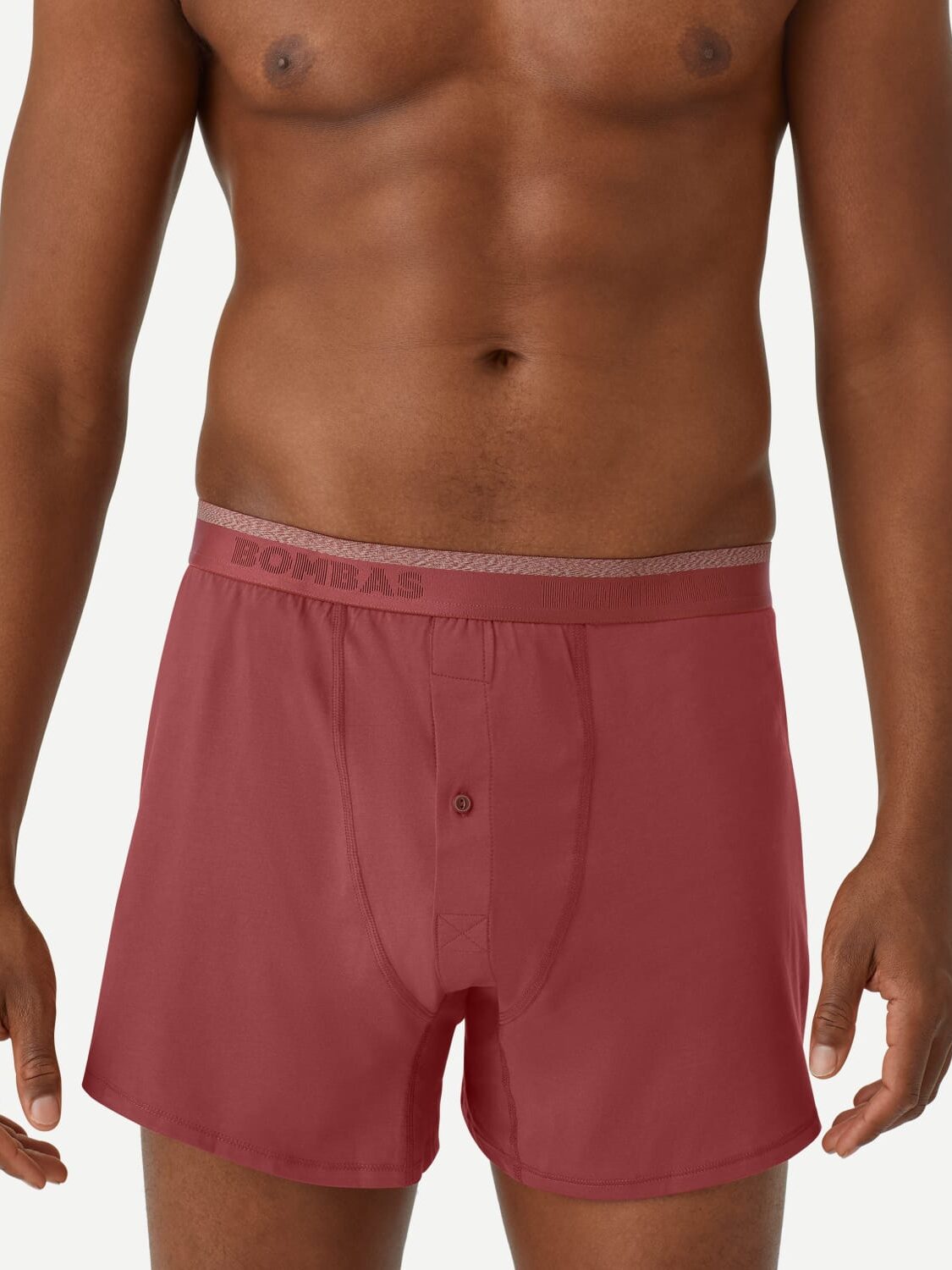 A person wearing red Bombas boxer shorts with a grey waistband is standing against a plain white background. The shorts have a button fly.