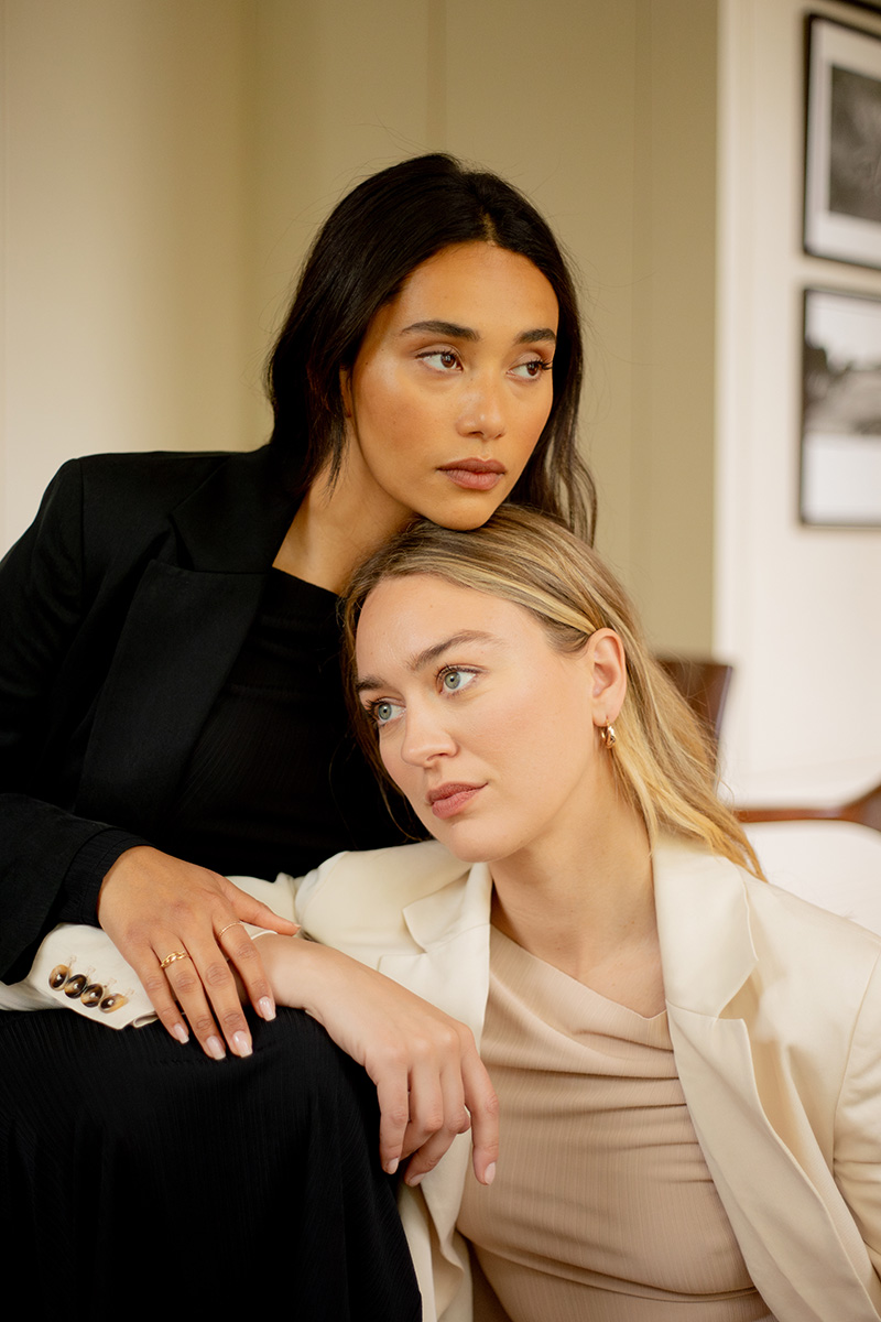 Two women sit closely; one in a black outfit leaning on the other seated in a white blazer. Both have serious expressions and look away from the camera.