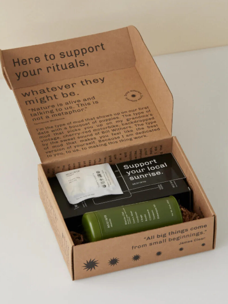 An open cardboard box reveals three items inside: a small white packet, a black box labeled "Support your local sunrise," and a green bottle. The box lid contains motivational text and designs.