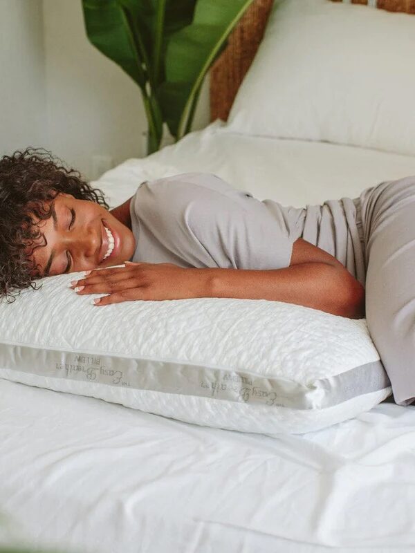 A person wearing a light-colored outfit is lying on a bed, smiling and resting their head on a white pillow. The background includes green plants and white bedding.