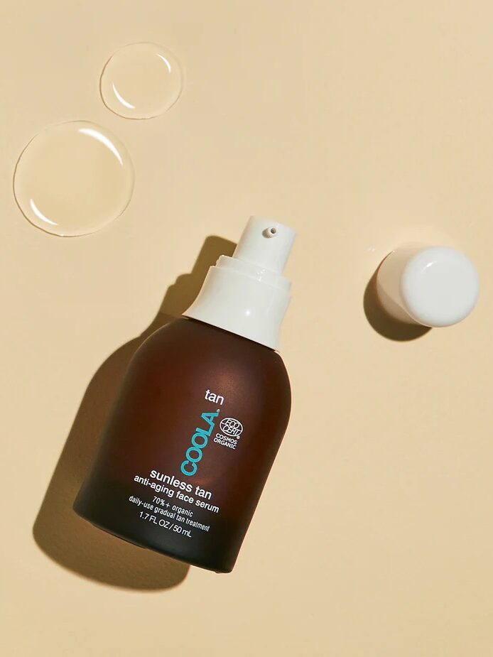 Brown bottle of Coola Sunless Tan Anti-Aging Face Serum with detached white cap and clear liquid droplets on a beige background.