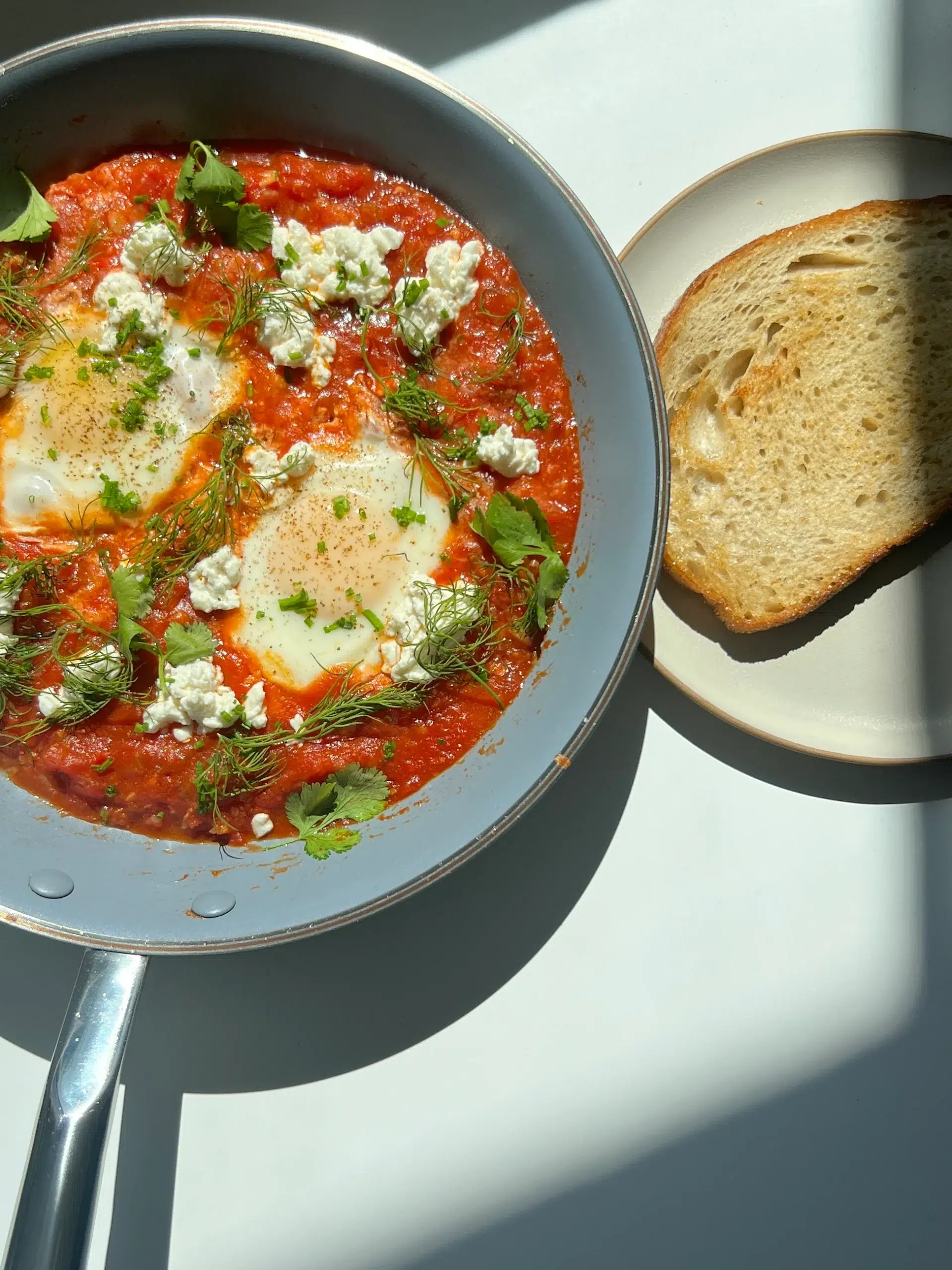 A pan containing shakshuka with poached eggs, tomato sauce, herbs, and cheese sits next to a slice of toasted bread on a plate under sunlight.