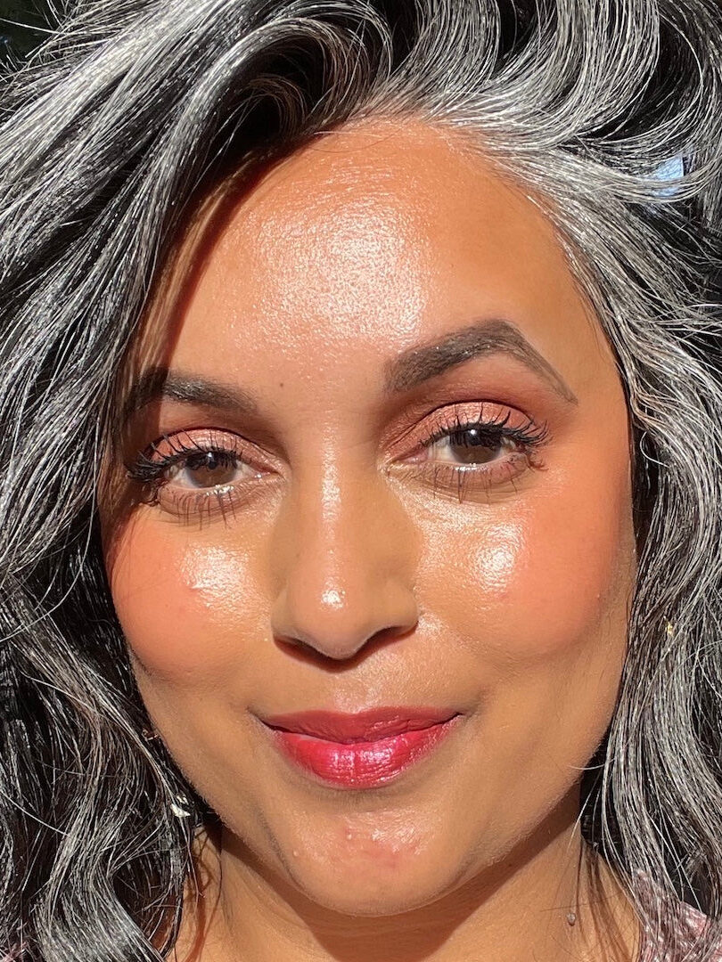 Close-up of a person with silver-streaked hair, wearing makeup, and smiling confidently in natural sunlight.