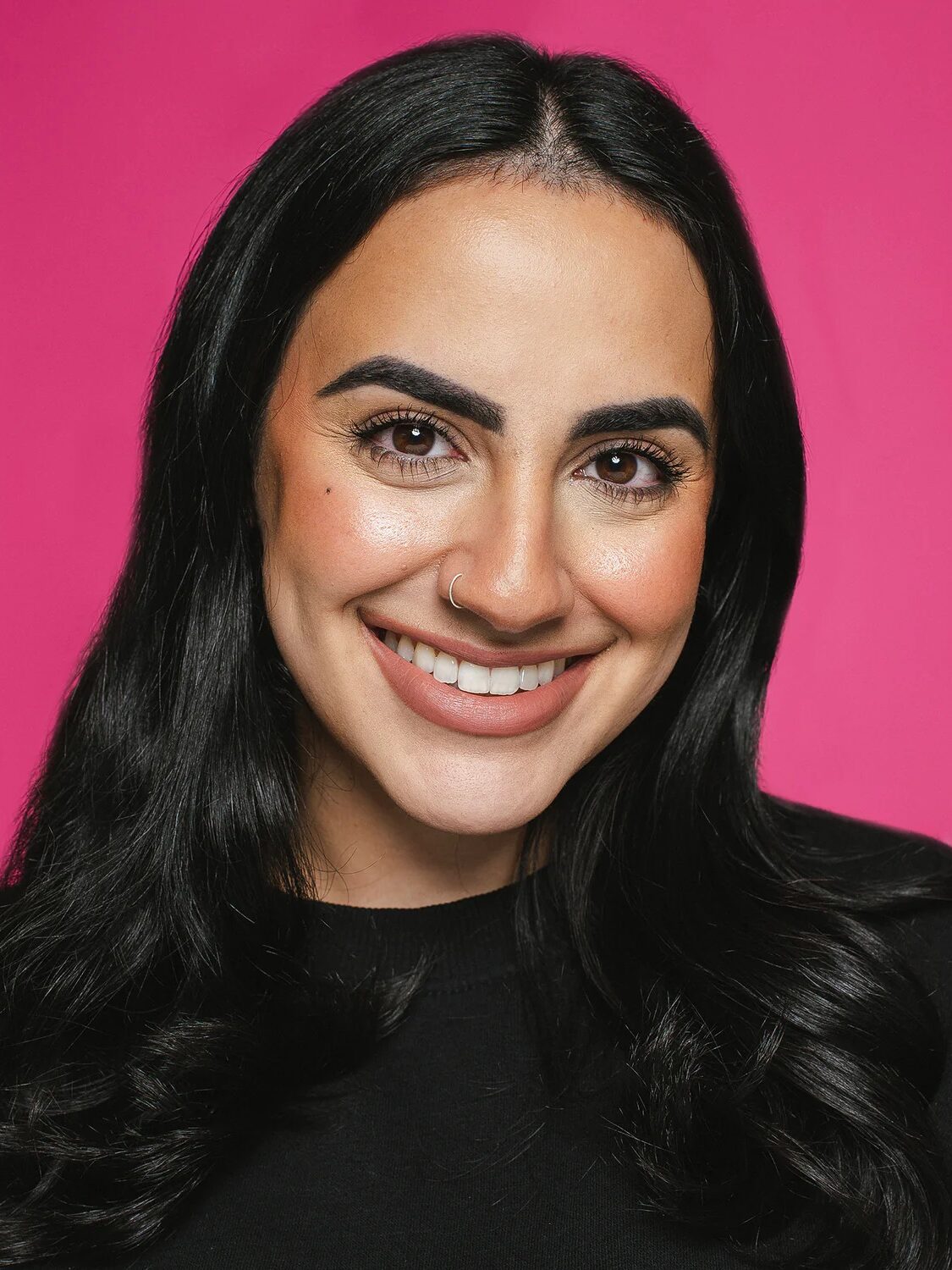 Woman with long black hair, wearing a nose ring and black top, smiling against a bright pink background.