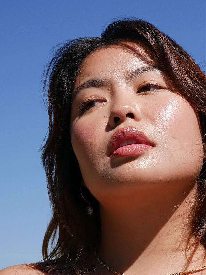 Close-up of a woman looking up against a clear blue sky, with sunlight illuminating her face. She has medium-length dark hair and is wearing a single pearl earring.