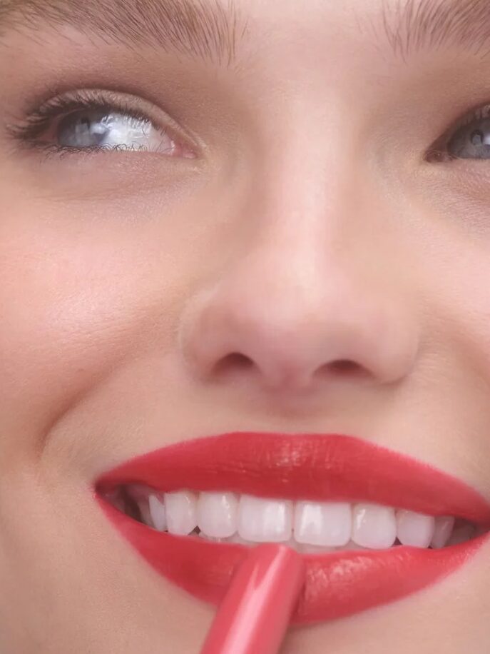 Close-up of a person applying pinkish-red lipstick to their smile, with a light complexion and smooth skin. The name "AUDREY" is displayed in the bottom left corner.