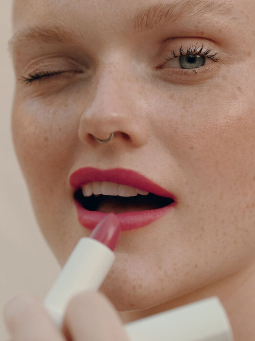 Close-up of a person with freckles applying pink lipstick, one eye closed. The person has fair skin, light blue eyes, and a small nose ring.