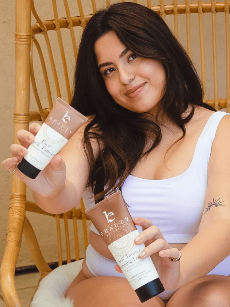 A woman in a white swimsuit sits on a rattan chair holding two skincare product tubes labeled "Self Tanner" and "Face Tanner.