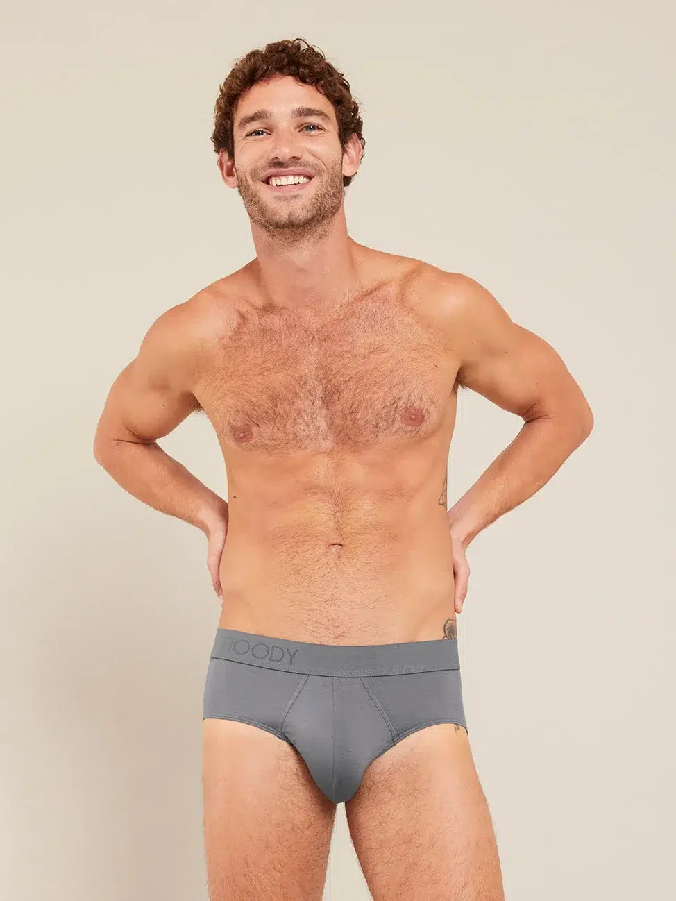 A man stands smiling with his hands on his hips, wearing grey briefs.