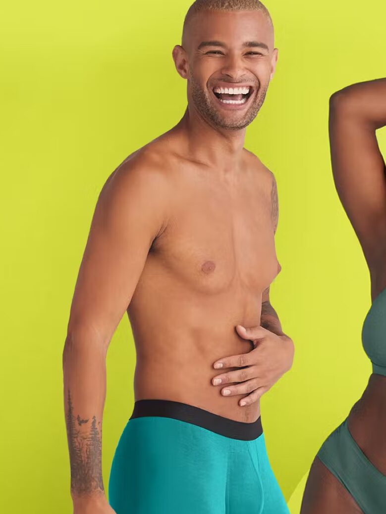 Two people in a cheerful pose against a lime green background, both wearing teal-colored underwear. The person on the left is smiling while holding their stomach, and the person on the right has an arm raised.