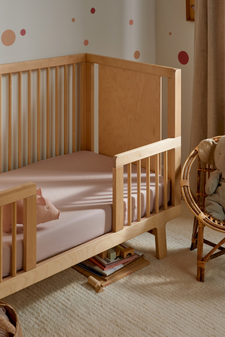 A wooden baby crib with pink bedding is placed next to a wicker chair. Books are stacked under the crib on a beige carpet. The wall has polka dot decorations.