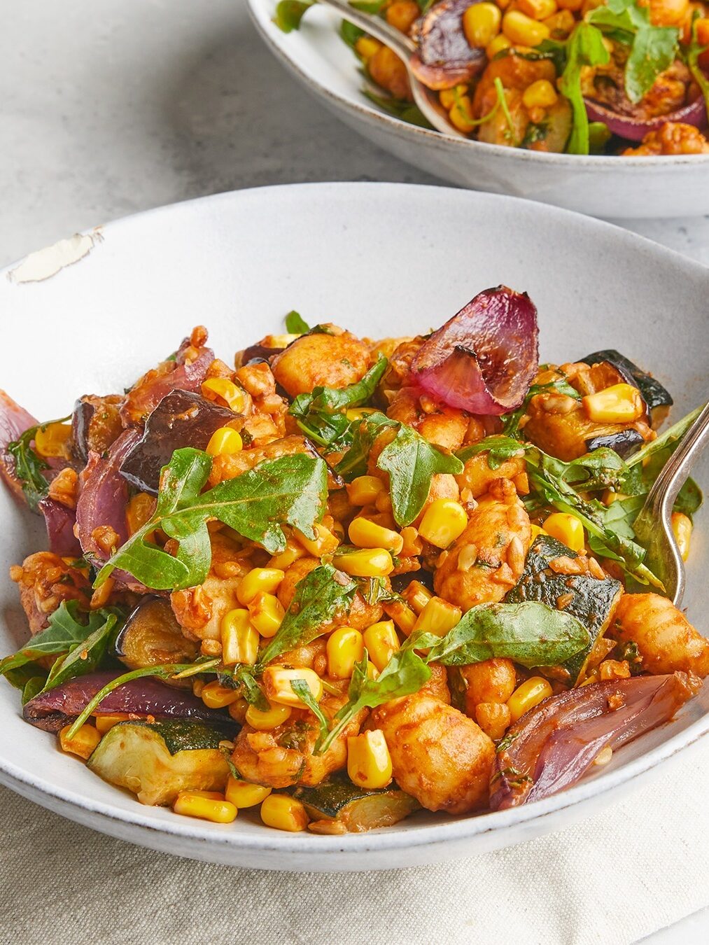 A white bowl filled with a mixed vegetable and gnocchi dish featuring corn, arugula, red onion, and other assorted vegetables, served on a light-colored surface. A fork is placed in the bowl.