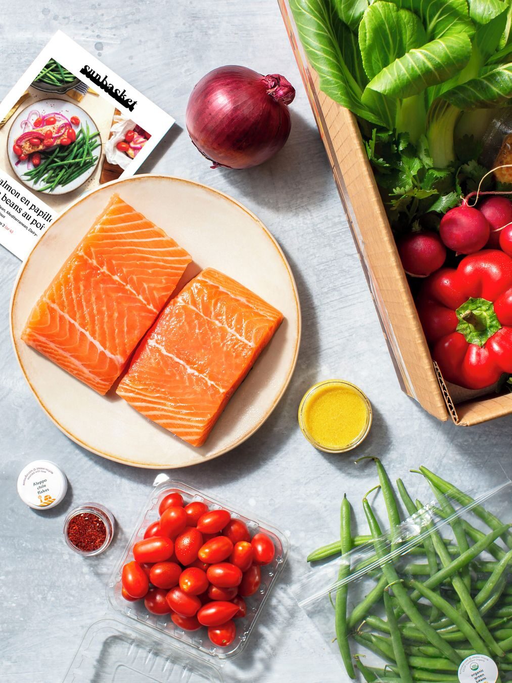 A variety of fresh ingredients including salmon, tomatoes, green beans, red onion, radishes, greens, and a jar of yellow sauce are arranged around a recipe card, with a box of vegetables nearby.