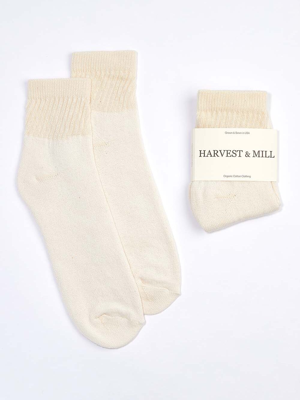 Two pairs of light beige organic cotton socks, one unfolded and one folded with a packaging label that reads "Harvest & Mill.