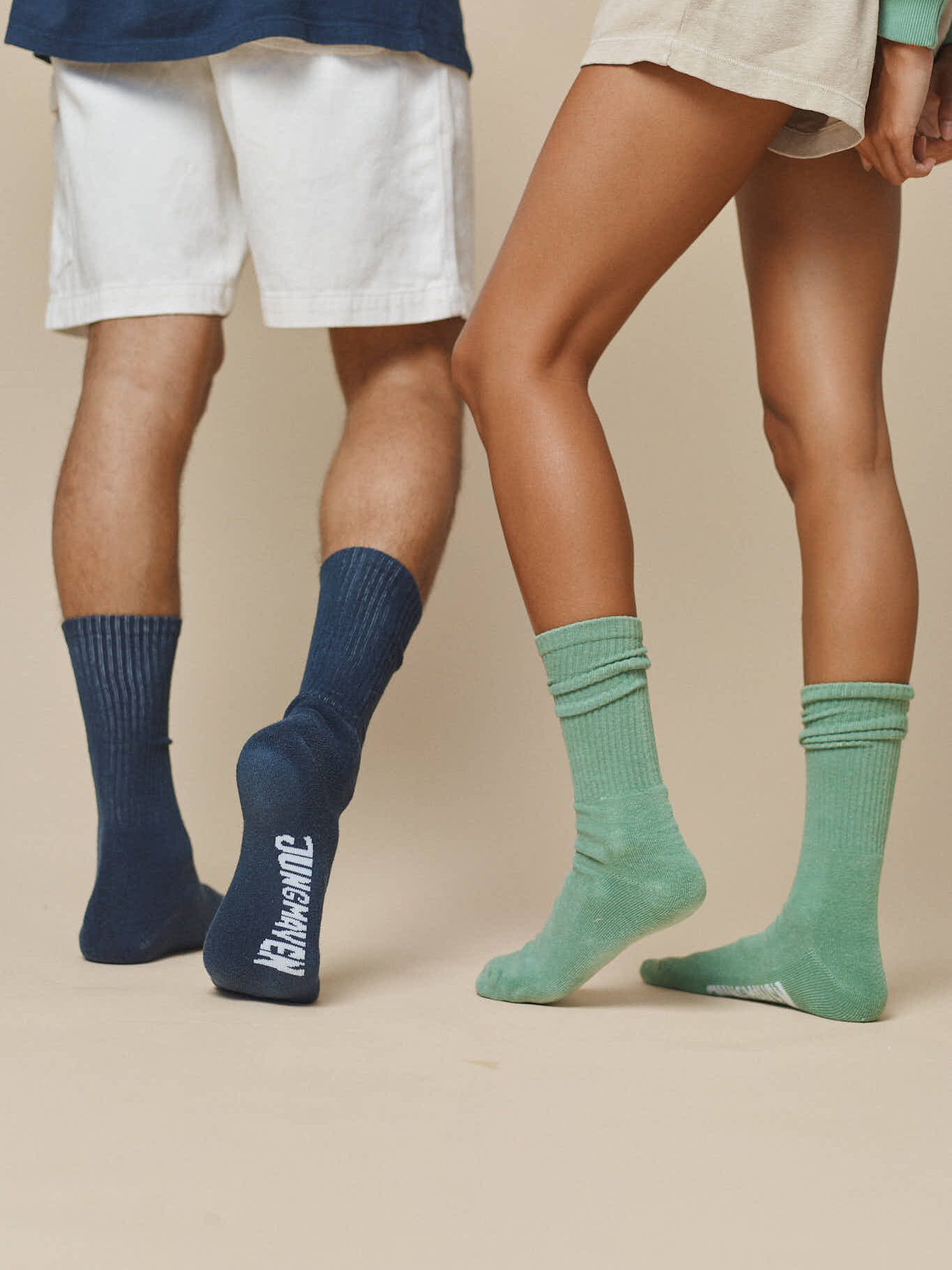 Two people showcasing organic calf-length socks, one wearing navy socks paired with white shorts, and the other wearing green socks paired with beige shorts. The background is plain beige.