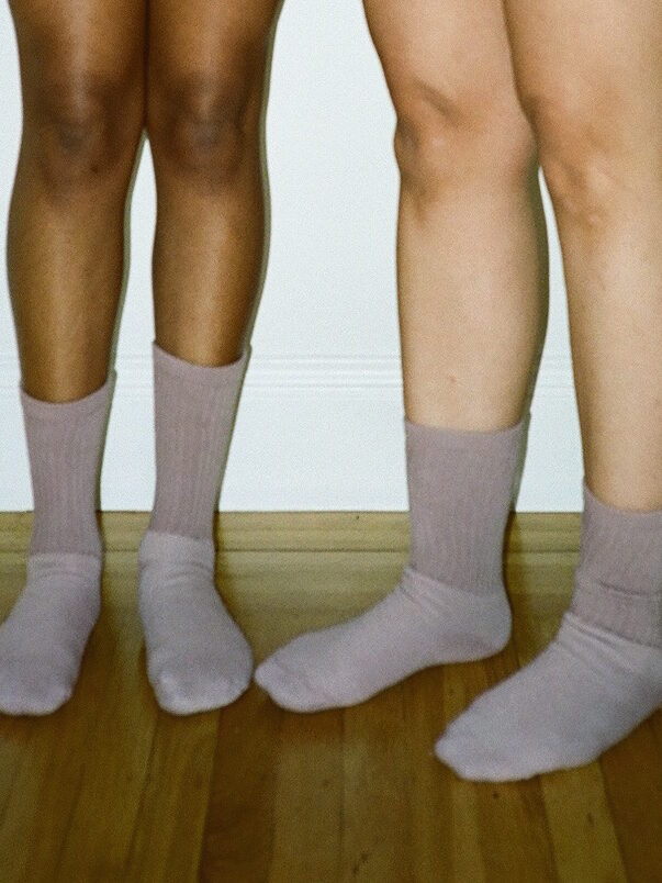 Two people are standing side by side on wooden floorboards, each wearing light purple organic socks.