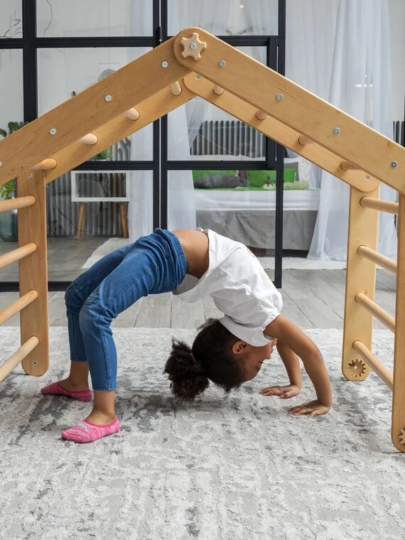 A young girl performs a backbend under a wooden play structure indoors. She is wearing blue jeans, a white shirt, and pink socks. The room has large windows and indoor plants.
