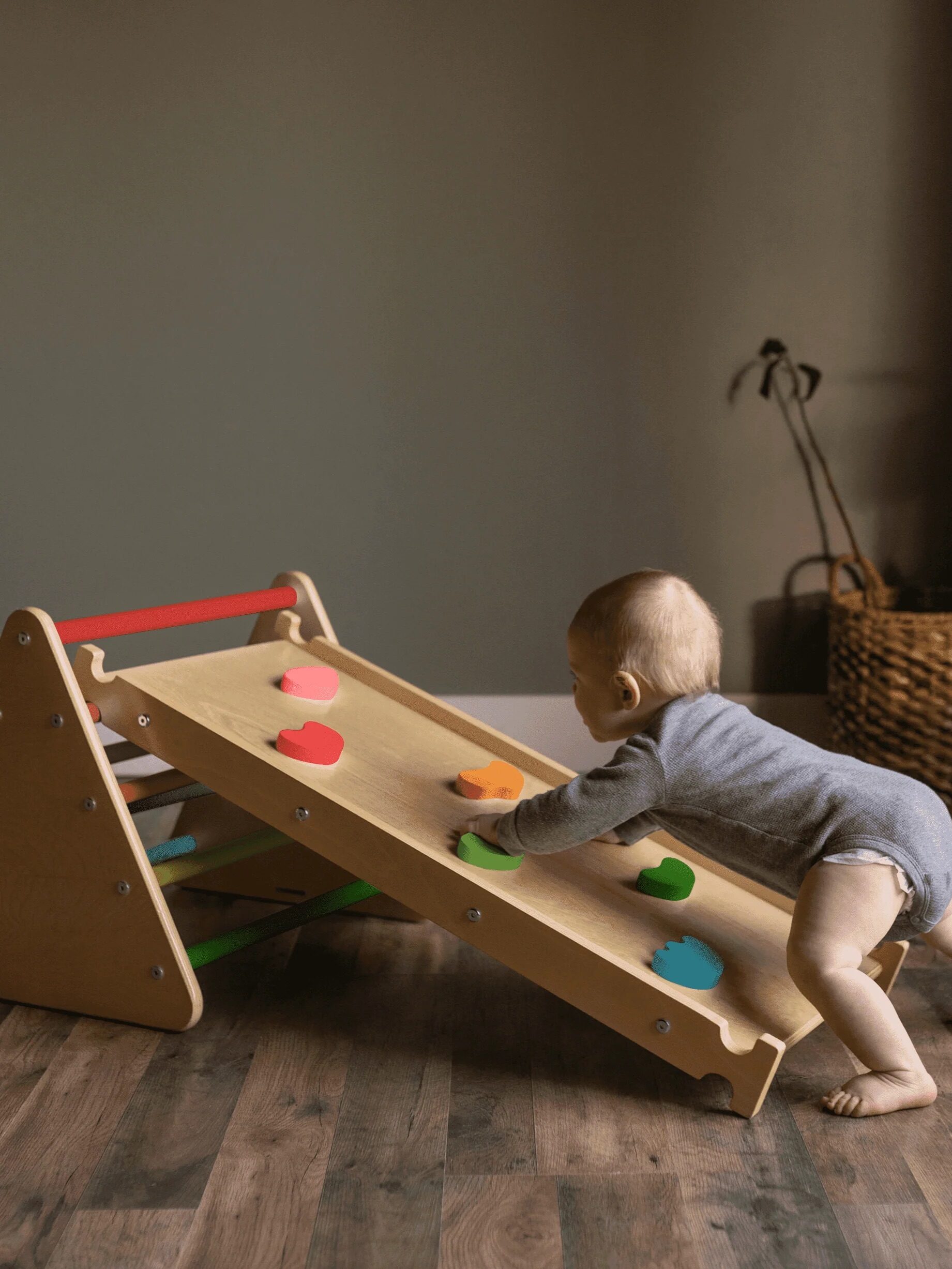 A baby in a grey outfit is playing with a wooden climbing ramp with multi-colored holds indoors on a wooden floor.