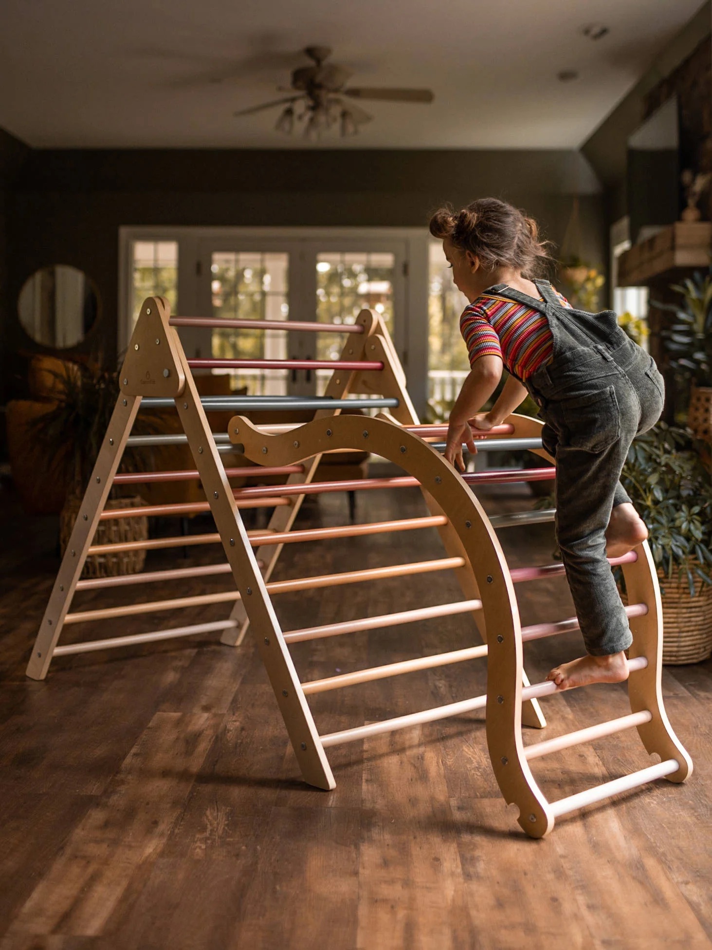 A child climbs on an indoor wooden play structure with a climbing ladder, in a well-lit room with wooden floors and large windows in the background.