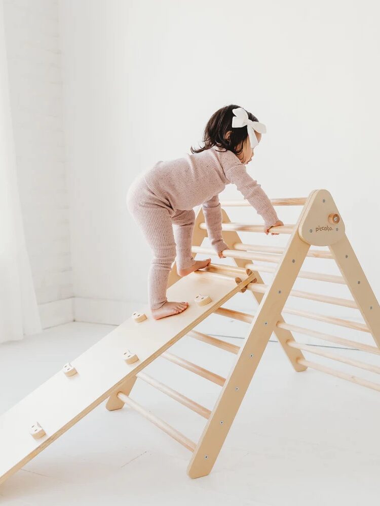 A toddler in a pink outfit with a white bow climbs a wooden indoor play structure in a bright room.