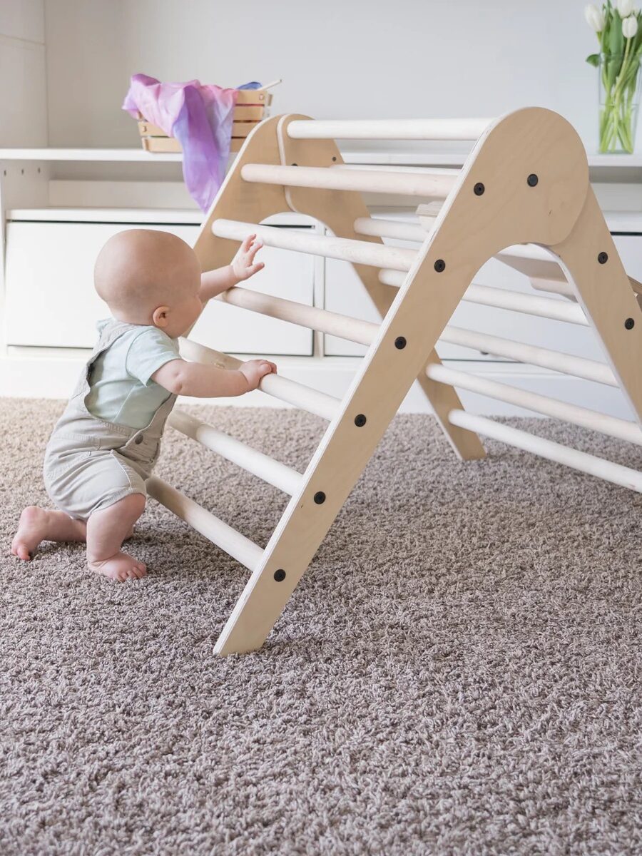 A baby wearing overalls is climbing a wooden triangular ladder toy in a carpeted room.