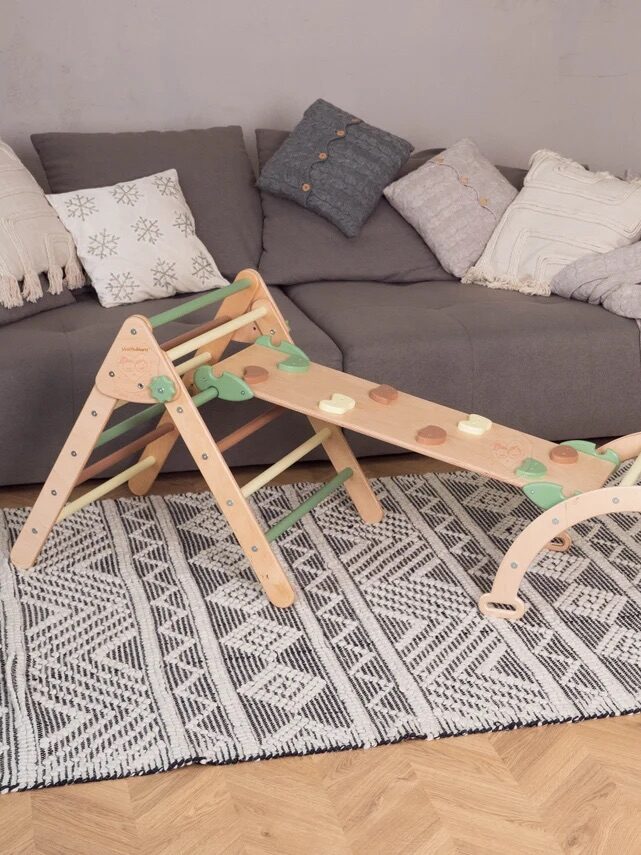 A wooden climbing structure for children is set up in a living room with a gray sofa, patterned rug, and pillows in the background. There is a mirror on the wall and wrapped gifts on the floor.