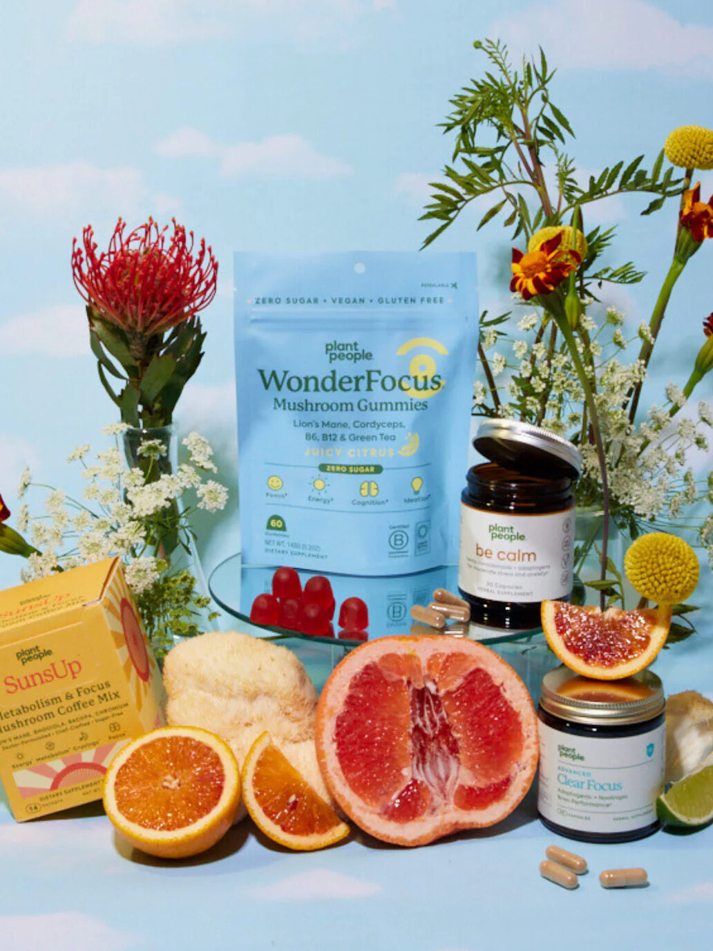 An arrangement of wellness products, including mushroom gummies, supplements, and sliced citrus fruits, set on a blue sky backdrop with flowers and greenery.