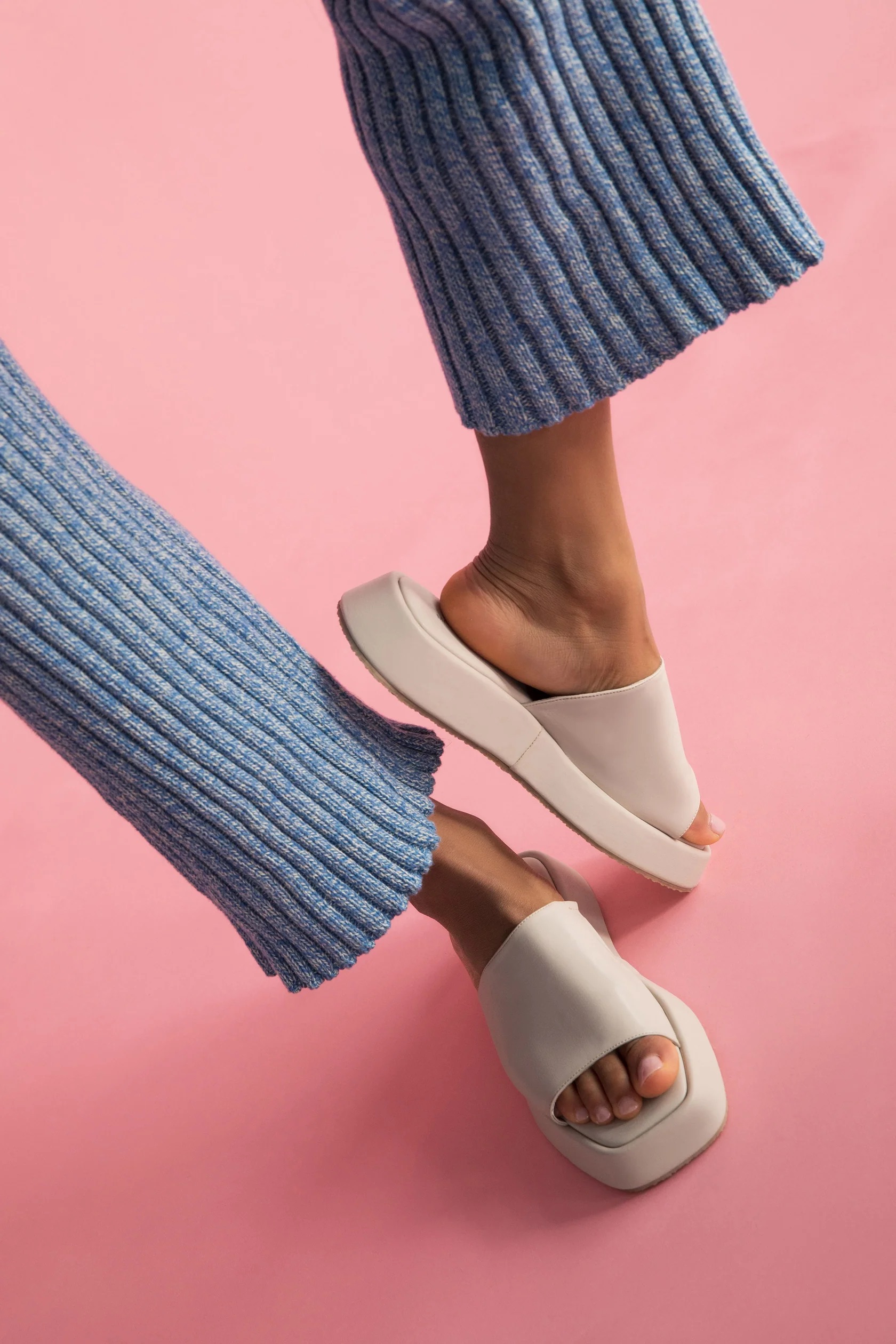 Close-up of a person wearing blue ribbed pants and beige platform slides with an open-toe design, positioned against a pink background.