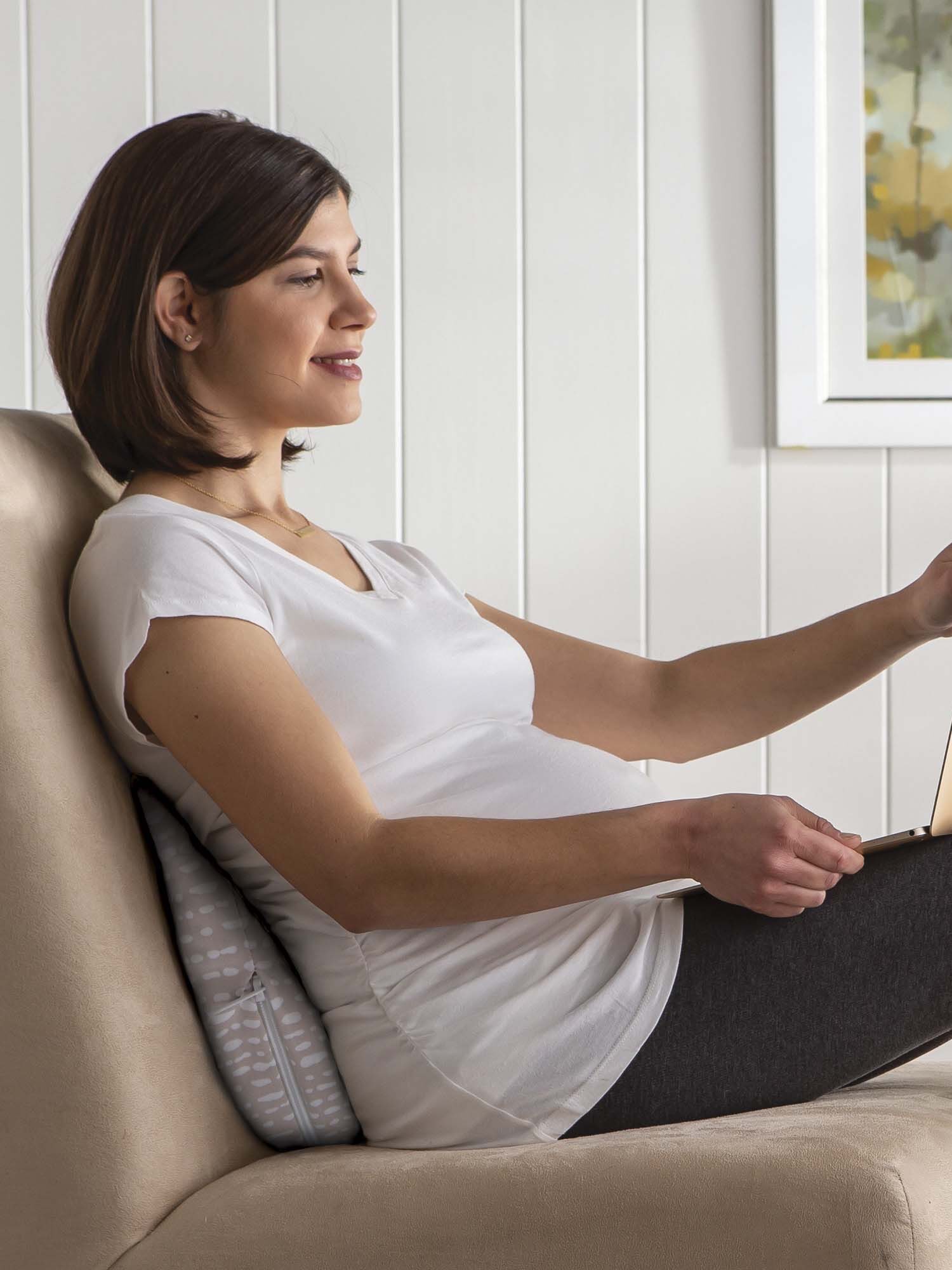 A pregnant woman in a white shirt and gray pants uses a laptop while reclining on a couch with a pillow for support. She appears to be working or browsing.