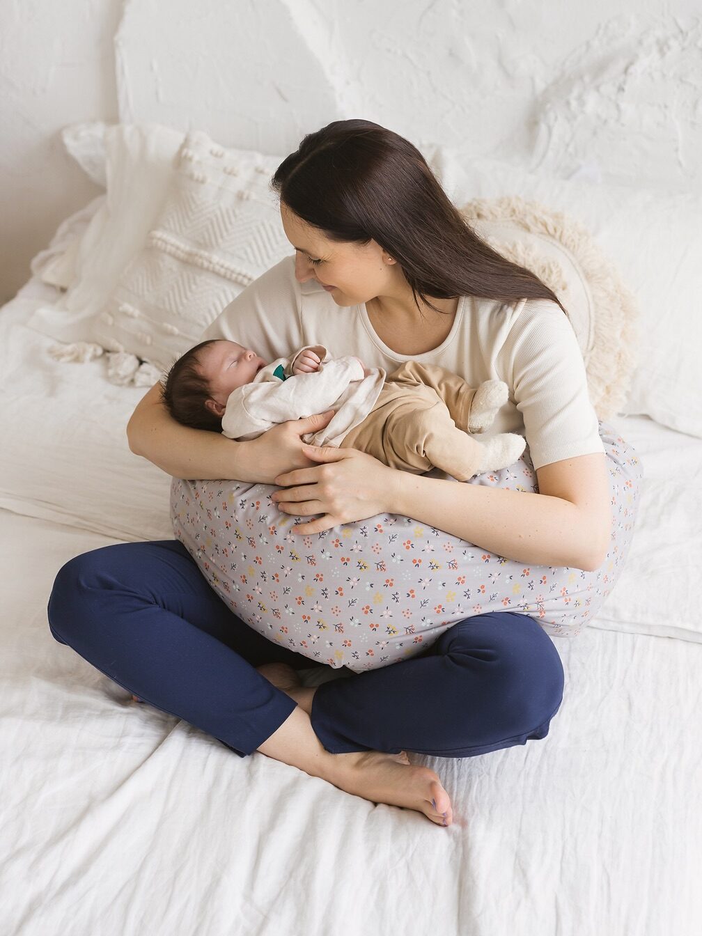 A woman is sitting cross-legged on a bed, holding a sleeping baby supported by a floral-patterned pillow. Both are surrounded by white bedding.