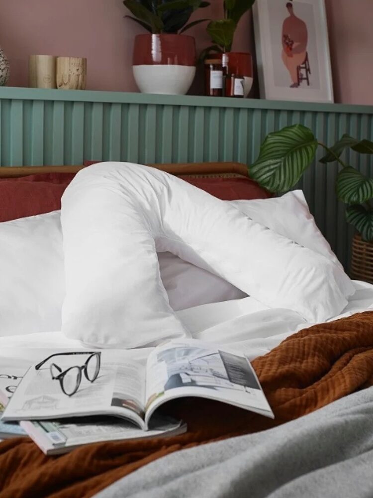 A white V-shaped pillow on a neatly made bed with an open magazine, a pair of reading glasses, and a rust-colored blanket in the foreground. Decorative items and plants are in the background.