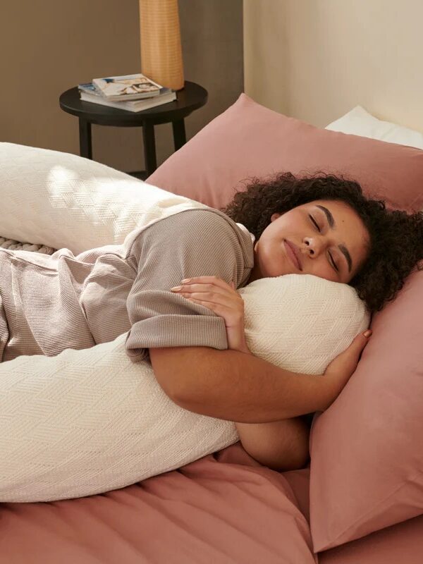 A person with curly hair is sleeping on a bed, hugging a large white body pillow. The bed has pink sheets and beige pillows. There is a small table in the background with a lamp.