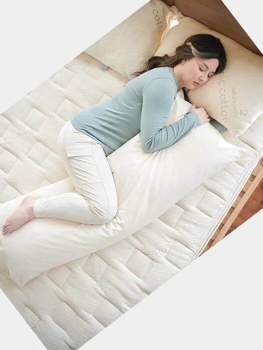 Person in a light blue top and white pants is sleeping on a bed, hugging a long body pillow.