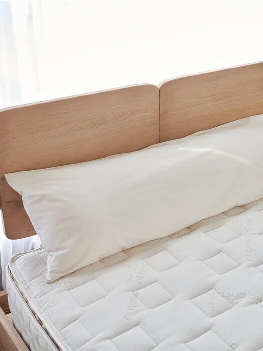 A neatly made bed with a wooden headboard, a white mattress, and a long white pillow. Sunlight streams through curtains in the background. There is a small round table with a drink beside the bed.