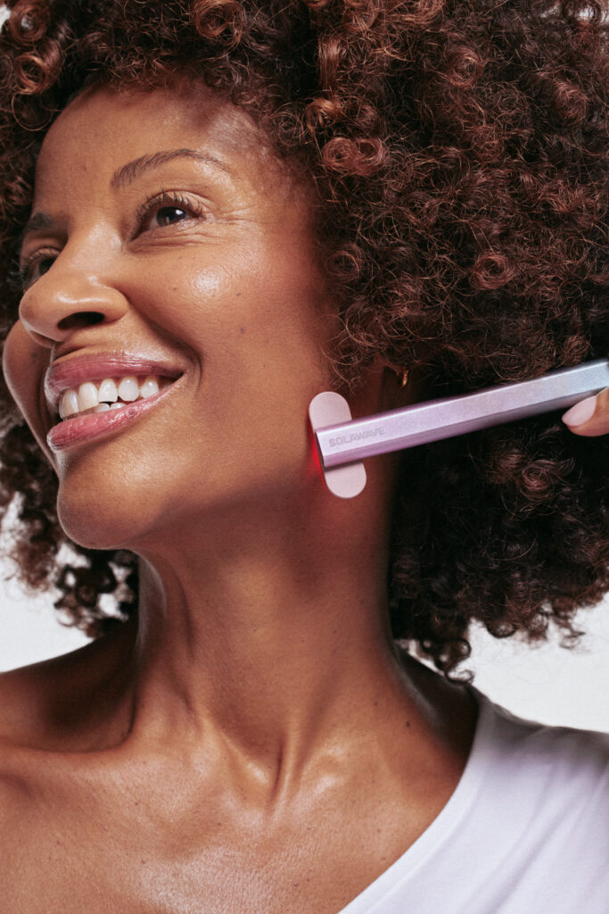 A smiling person with curly hair uses a skincare device on their chin, wearing a white top.