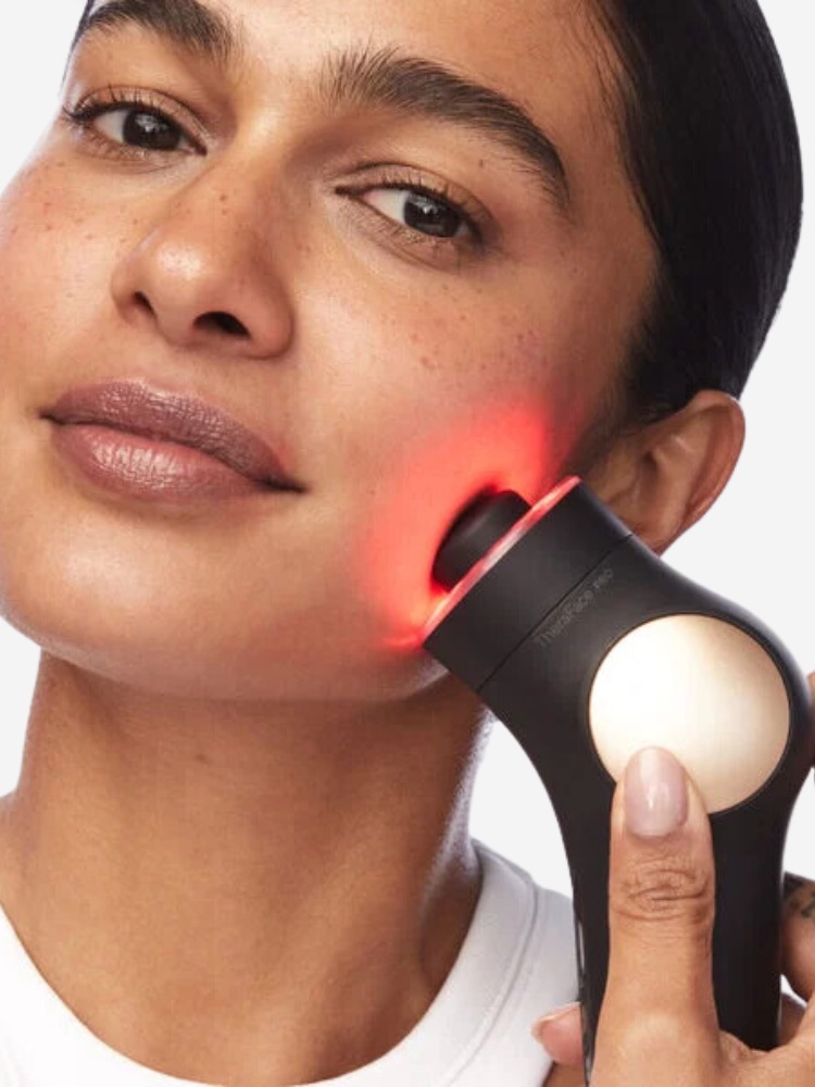 A person uses a handheld, black beauty device emitting red light on their cheek.