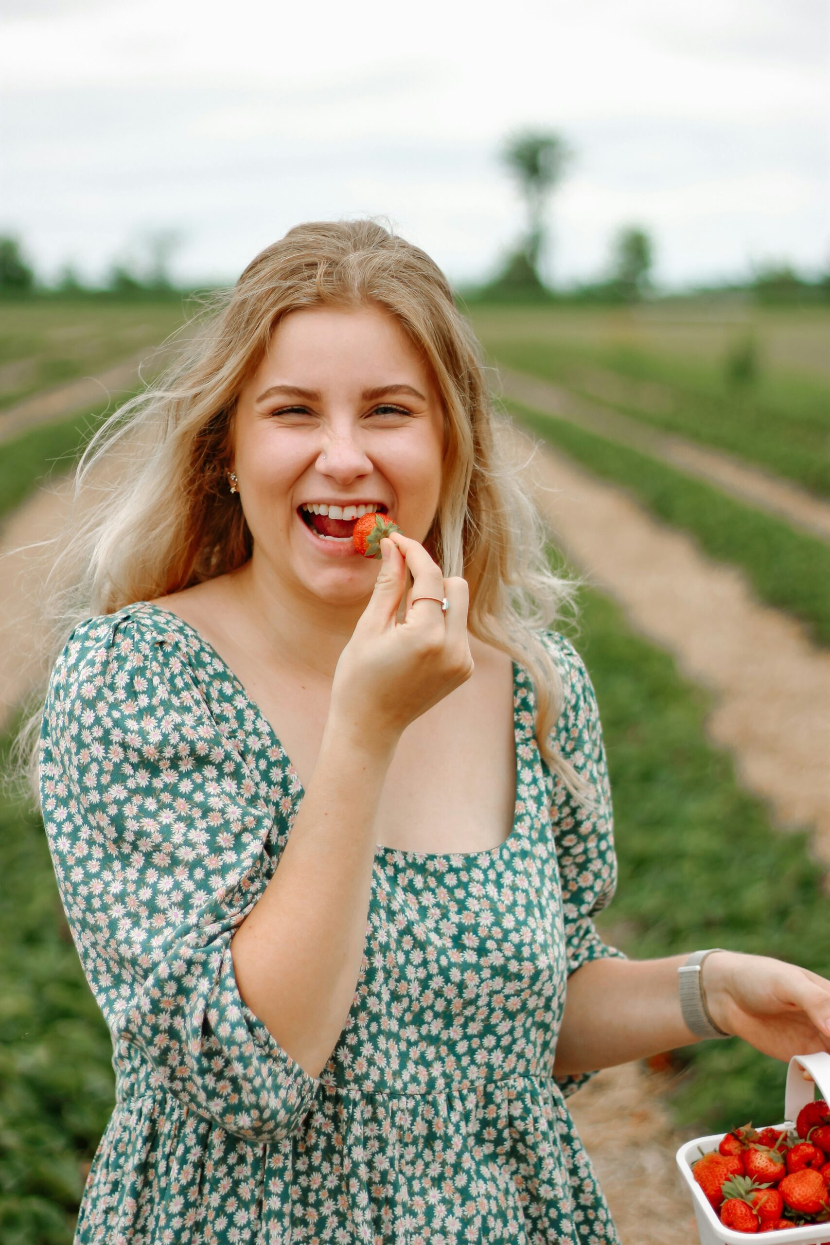 A person with long blonde hair in a green floral dress smiles and holds a strawberry to their mouth while standing in a strawberry field. They have a container of strawberries in their other hand.