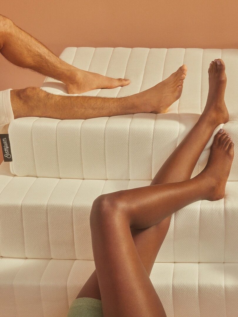 Two pairs of legs, one male and one female, rest on a stack of three white mattresses against a peach-colored background.