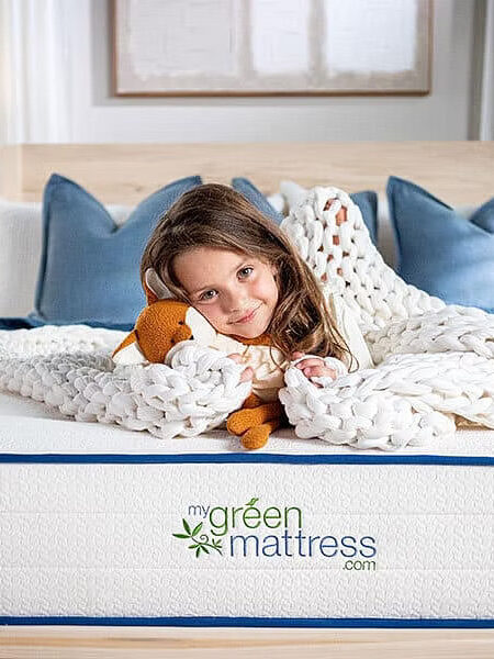 A young girl lies on a bed with a chunky white knit blanket and a stuffed toy. The bed features a “my green mattress” logo. Two lit table lamps and pillows are in the background.